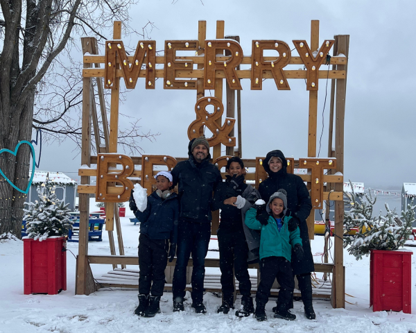 Family with three kids, dad, mom standing in the snow in front of a sign that says "merry"