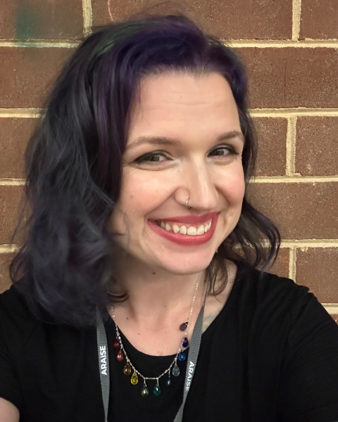 A woman with black short curly hair and a purple streak at the front. She is wearing a black t-shirt, a colourful necklace and a black lanyard.