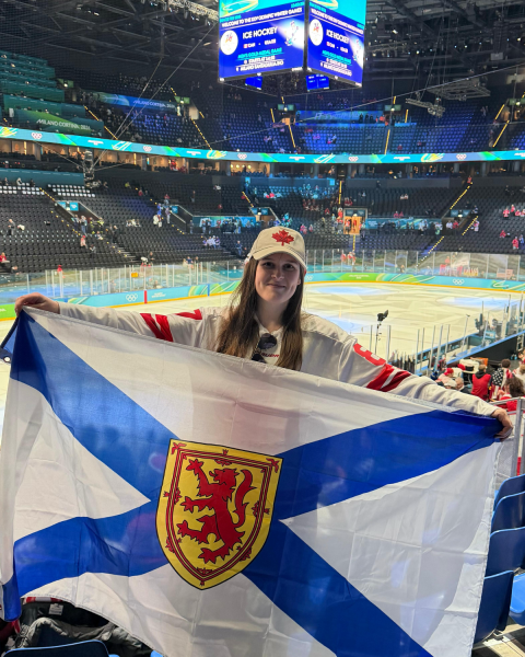 A photo of a woman in front of an ice rink. She is wearing a white hat with a red maple leaf on it. She has a red and white jersey on and she is holding a flag with a blue cross and a red and yellow crest in the front.