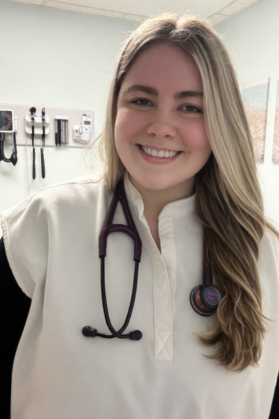 Photograph of woman wearing white scrub top and a stethoscope around her neck, smiling with long blonde hair. Behind her, the wall of a medical office can be seen with a few medical supplies hanging up. 