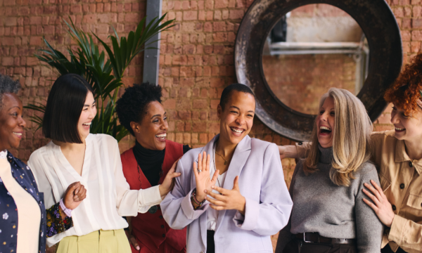 Photo of 6 women smiling and looking at each other. Mix of black and white women in a room with brown brick behind them, a plant and a round dark framed mirror.