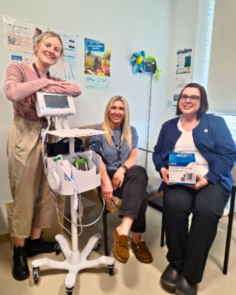 Picture of 3 women. Woman on the left is leaning over an automatic blood pressure machine, woman in the middle is getting her blood pressure taken and there is a woman in scrubs on the right side of the picture holding a box. They are all smiling largely. 
