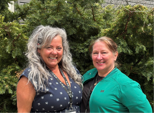 Two women stand side-by-side in front of a green leafy bush, smiling at the camera.