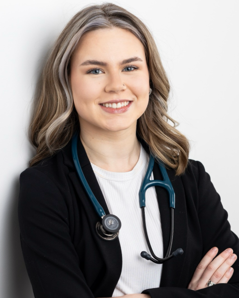 A photo of a woman with blonde curly hair. She is wearing a black blazer over a white shirt. She has a stethoscope around her neck.