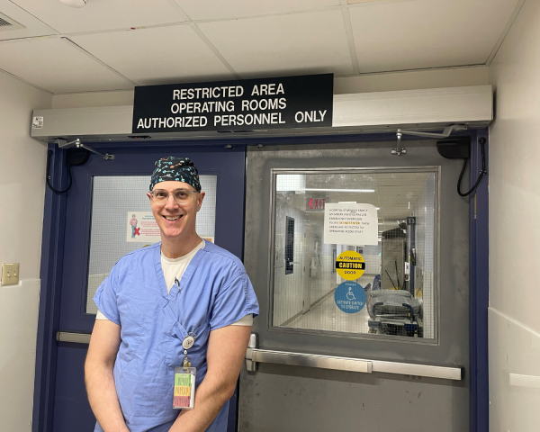 Picture of a man in blue scrubs and scrub hat standing in front of operating room doors. There is a sign above his head that reads "Restricted area operating rooms authorized personnel only."