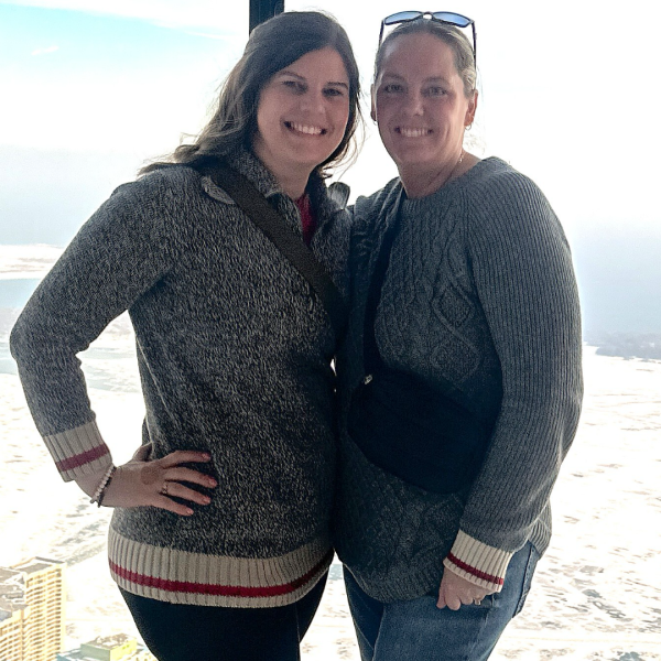 Two women are standing close together and smiling warmly for the camera. The background opens out into a bright cityscape and water view. 