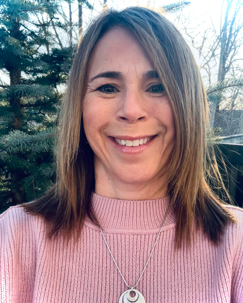 Photo of a woman with dark brown shoulder length hair smiling largely. Has a pink sweater and a necklace with a large pendant around her neck. The photo is taken outside with trees in the background.