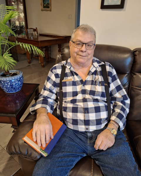 Man with glasses and short hair sits in a chair wearing a plaid shirt, suspenders and jeans.