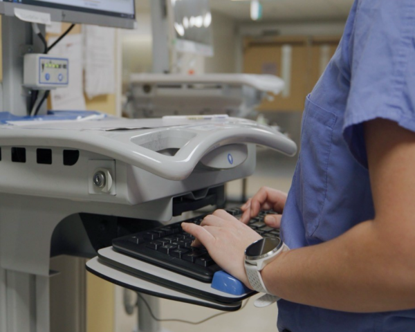 Image of a healthcare worker typing on a keyboard. The healthcare is standing using a WOW (workstation on wheels).