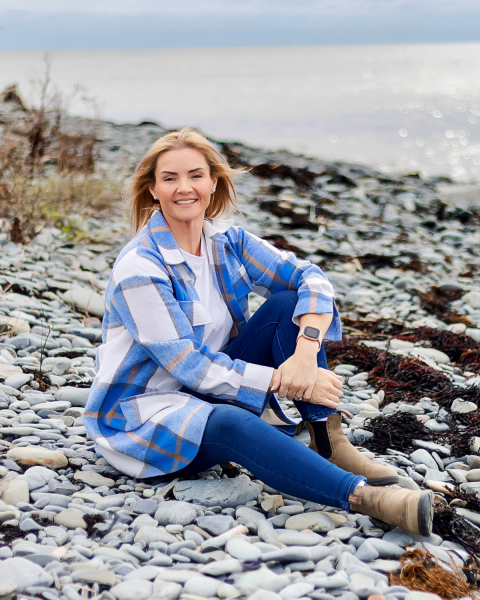 A photo of a woman at the beach city on the rocks. She has blonde hair and she is wearing a blue and white plaid jacket over a white shirt, jeans and boots.