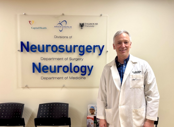 Man in a white lab coat stands in front of a sign on a wall inside a healthcare facility that says "Neurosurgery" and "Neurology".
