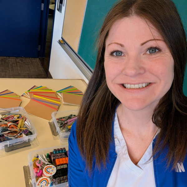 A smiling woman fills the right side of the frame, taking a cheerful selfie in what looks like a classroom or creative workspace. Behind her, a tabletop is neatly arranged with craft materials. 