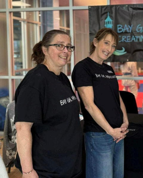 Two women indoors, standing side by side, both wearing black t-shirts with the text “Bay Gals” printed on them.  