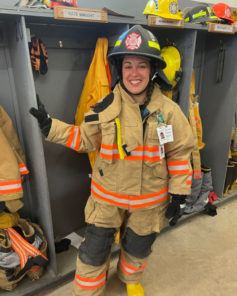 Image of a female firefighter in full gear standing next to a open closet with firefighter gear hanging up.