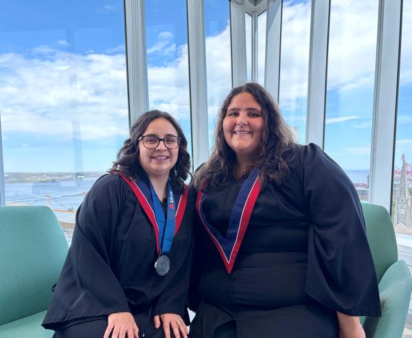 photo of two women in graduation regalia sitting in front of a window