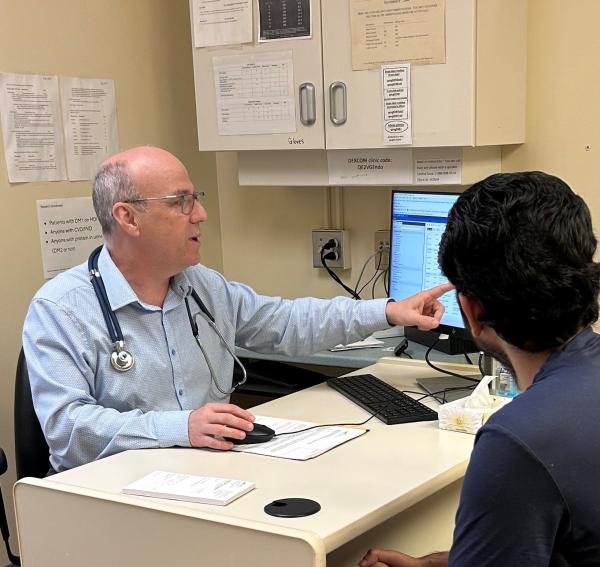 A man wearing a stethoscope and blue shirt sits at a desk across from another person and is pointing to a computer screen.