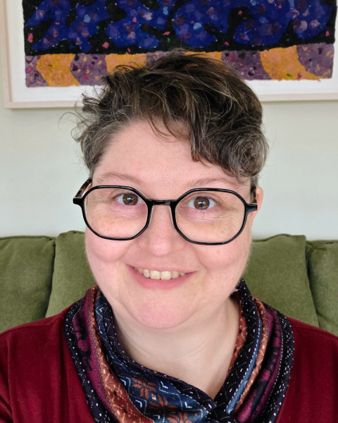 Close up image of a woman with dark curly short hair, wearing glasses and smiling. She is wearing a red top and multicolour scarf.