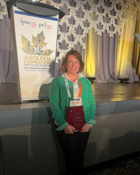 A woman with blonde short hair is wearing a green sweater is smiling at the camera holding an award.