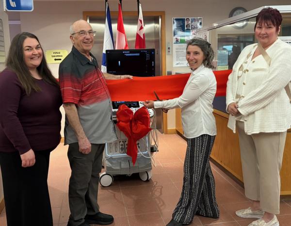 Four people pose with an ultrasound machine with a ribbon tied around it to celebrate its arrival at a local hospital.