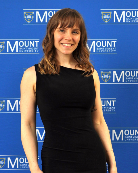 A smiling woman is standing in front of a blue backdrop covered with the Mount Saint Vincent University logo and text. 