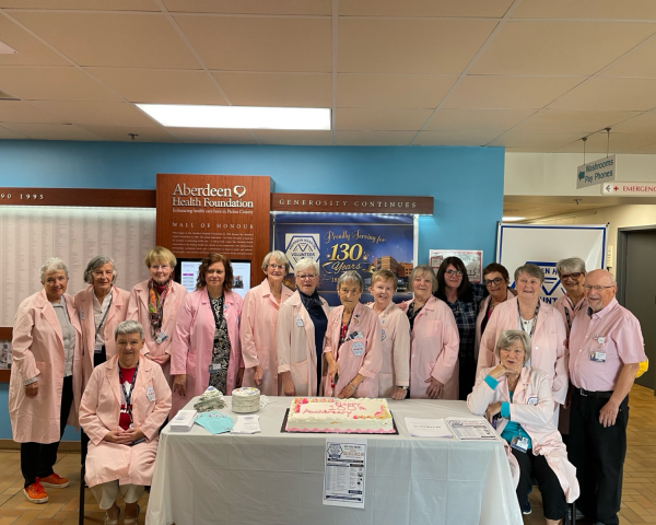 Image of a large group of women and one male in the picture. All people are wearing light pink lab style long jackets. There are 2 women sitting and the rest are standing - all around a table with a white table cloth and a cake sitting on top.