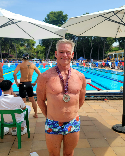 Close up image of a man wearing bright coloured swim trunks. He is standing wearing a medal around his neck and is smiling. In the background are umbrellas and a pool.