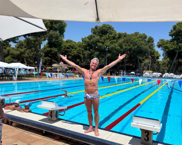 Photo of a man wearing swimming trunks standing at the end of a pool with his arms opened wide. He is wearing a medal around his neck.