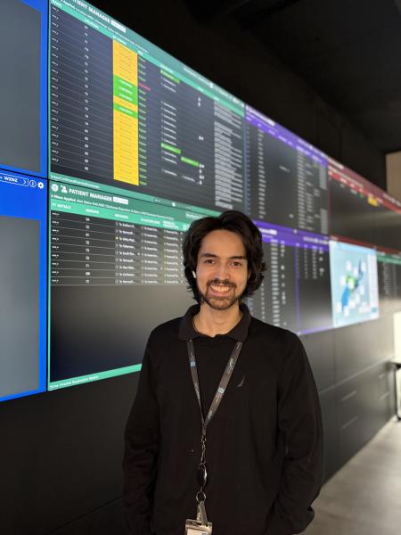 Man standing in front of digital screens wearing a black shirt, lanyard, and has dark hair and a beard.