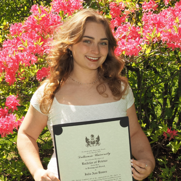 In an outdoor setting, a smiling young woman stands in bright sunlight while holding a framed diploma in front of her. 
