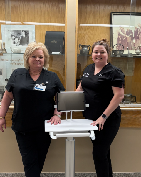 Two healthcare workers in black scrubs are standing inside a building with glass display cabinets behind them. They are standing with a portable computer between them.