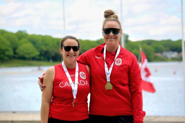 Two women on a podium wearing sunglasses, red shirts, and both have a medal around their neck. 