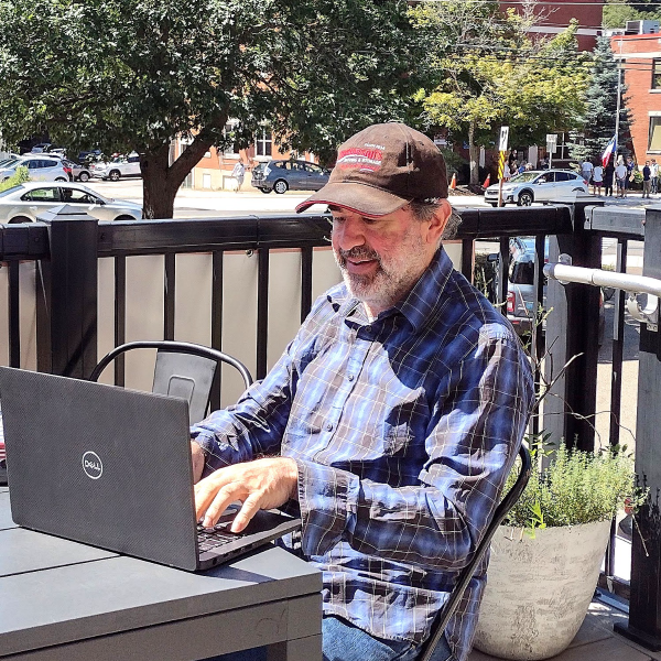 A smiling man sits outdoors at a table and is focused on using a laptop. He has both hands resting on the keyboard.  
