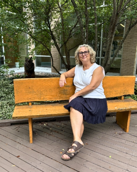 Debbie Burris (woman wearing a white top and navy skirt, short light hair and glasses. Sandals on her feet) sitting on a bench in the Sheila Gloade Memorial Healing Garden.