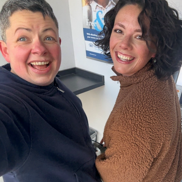 A close up image of two women smiling largely together. One woman on the left has short hair and is grey in colour, the woman on the right has shoulder length dark brown hair.