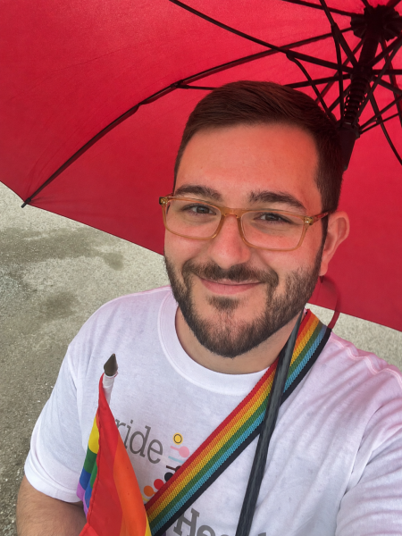 Person with short hair, glasses and wearing white shirt holding a rainbow flag and standing under a red umbrella, smiling at the camera.