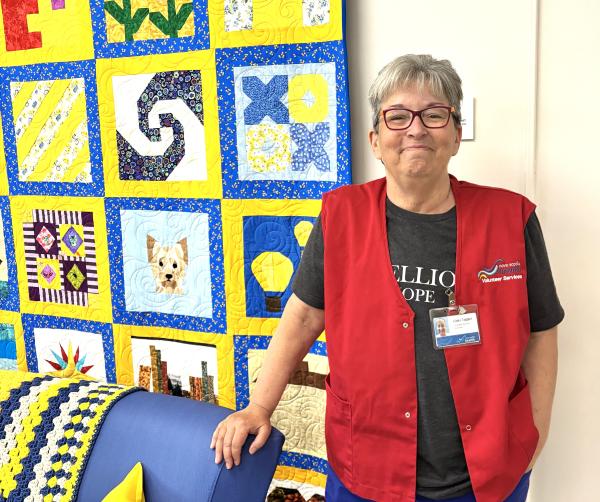A smiling woman stands indoors beside a brightly colored quilt display, resting one hand on the back of a blue couch. 
