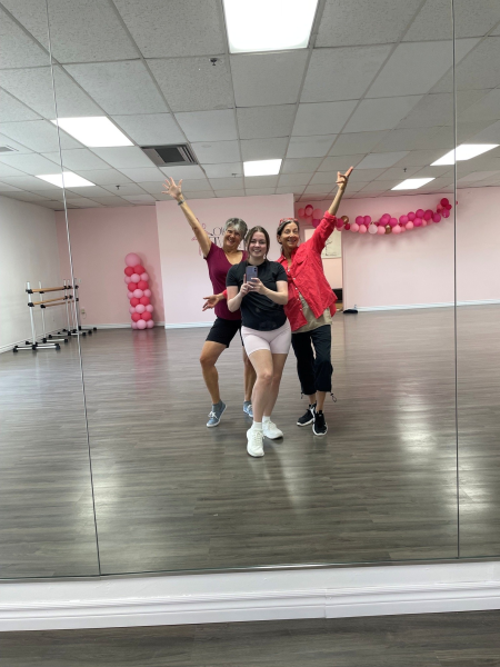 Three women in a dance studio, with arms extended, posing for a photo in front of a mirror. 
