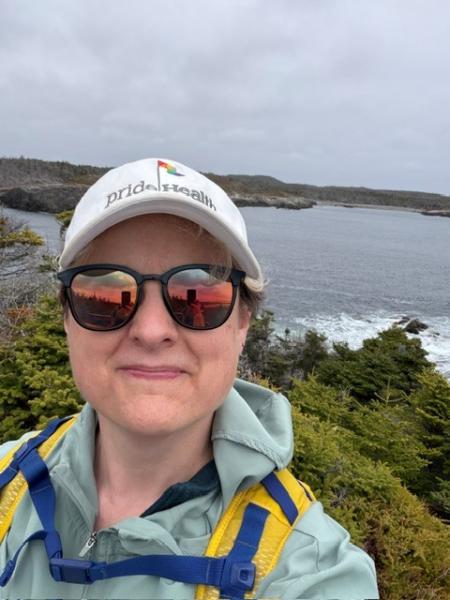 A person stands outdoors wearing a ball cap, sunglasses and a raincoat, smiling at the camera.