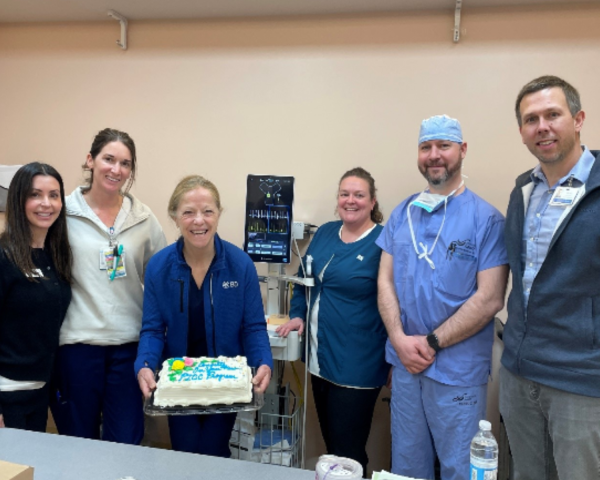 Six healthcare colleagues surround a PICC machine with a cake. 