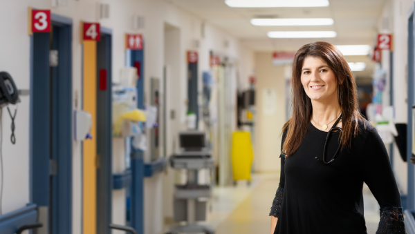 A healthcare professional wearing a stethoscope stands in a hospital hallway, smiling at the camera, with treatment rooms and medical equipment visible in the background.