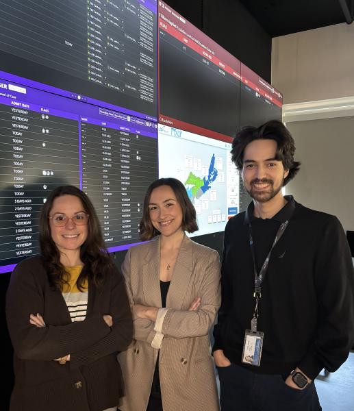 Three people stand in front of a series of digital screens, smiling at the camera.