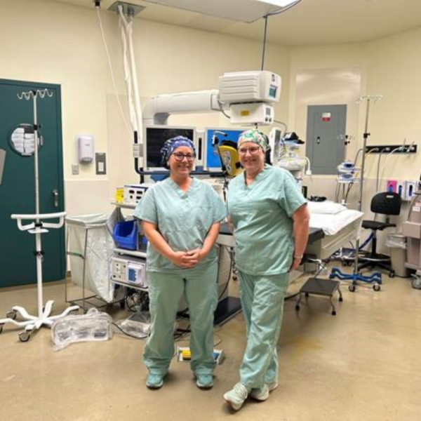 Two people stand side-by-side in an operating room wearing hospital scrubs.