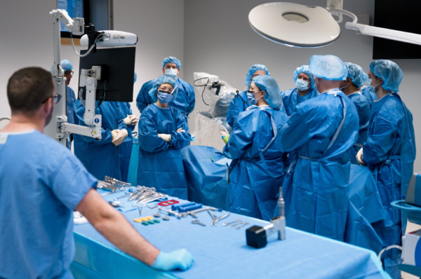 A group of people wearing blue surgical gowns stand around a hospital bed in a surgery room.