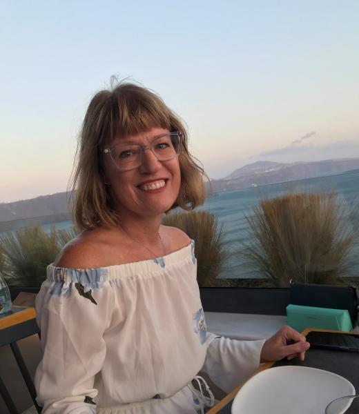 A woman wearing a white and blue dress poses, seated, with mountains in background.