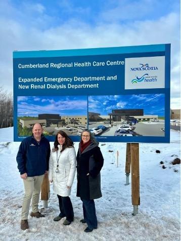 An image of 3 people standing outside with winter jackets on and there is snow on the ground. They are taking the picture in front of a sign that indicates a new hospital project underway.