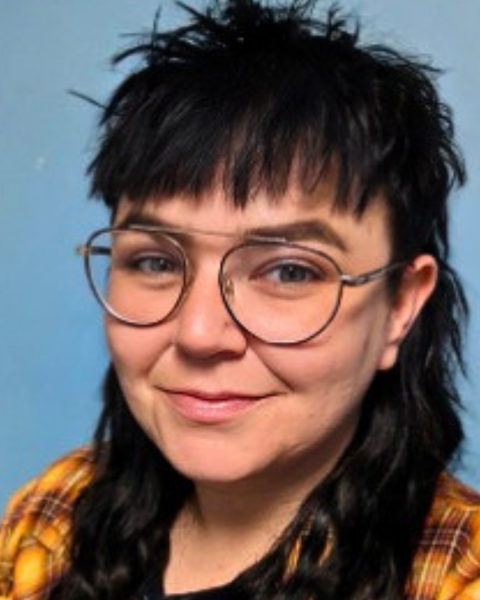 Close up image of a woman with long black hair and bangs. Wearing glasses and a yellow plaid shirt. There is a blue background behind her.