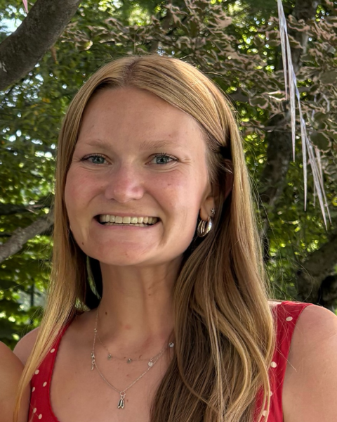 Close up image of a woman with long dirty blonde hair. Smiling largely. Standing outside with greenery behind her. She is wearing a short sleeve red polka dot top.