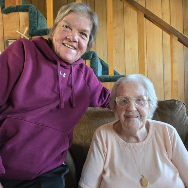 Close up image of two women, smiling largely at the camera. One woman on the left is standing next to an elderly lady on the right wearing glasses. The elderly lady is sitting down.