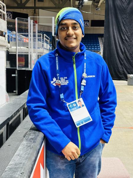 A man wearing a blue hospital scrub hat, blue jacket and a lanyard with his name tag is smiling at the camera.