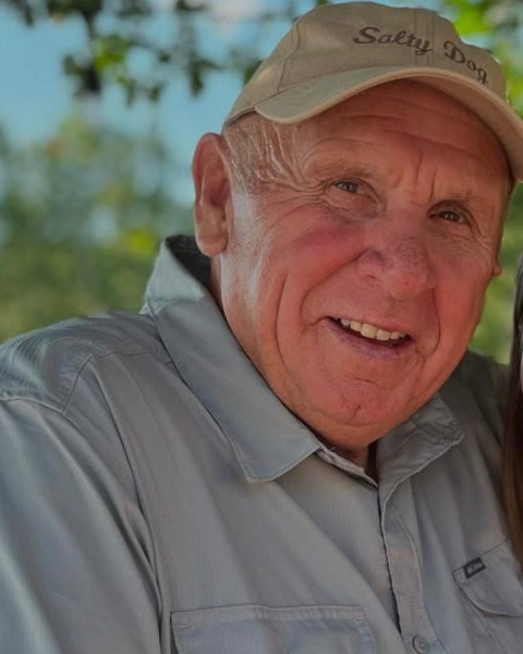 A man wearing a beige baseball cap and a gray collared shirt smiles gently at the camera, with soft greenery and blue sky in the background.